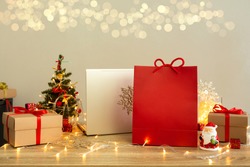 Shopping bag with mockup and laptop among gift boxes on wooden table with blurred lights background. Christmas shopping online concept.