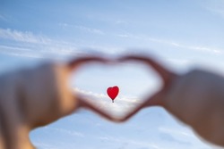 A heart from the hands on the background of the blue sky, a large balloon in the shape of a heart is visible in the frame of the fingers