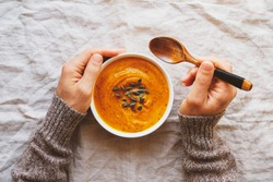 Male hands holding a bowl of hot pumpkin soup served with squash seeds on white tablecloth background, from above. Flat lay. Homemade autumn food. Popular Thanksgiving dish.