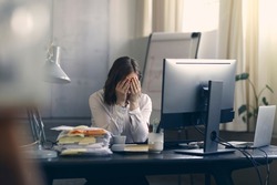 Very stressed business woman and female employee sitting in front of her computer with her hands in front of her eyes, feeling sad and depressed. Too much work - huge pile of paperwork at office table