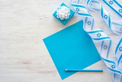 Blank notebook paper, blue pencil and Israeli symbols and items with national colours for Independence Day of Israel on white wooden background with copy space.