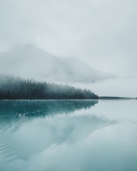 Thick, eerie fog creeping over pine trees with towering hazy mountains across the emerald water reflections of Emerald Lake  in Yoho National Park, BC, Canada.