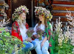Happy girls look at each other in large floral wreaths, Ukrainian national scarves on their shoulders with bouquets sitting. Background rural wooden house. Traditions in Ukraine. Summer concept.