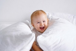 Laughing toddler having fun on bed with lots of pillows. A baby playing hide and seek.