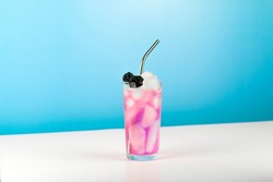 Summer refreshing pink cocktail with ice cube, metal straw and blackberry on blue background.
