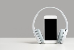 Smartphone mockup display with grey wireless headphones on white desk gray background. Copy space. Audio technology apps, music podcasts books