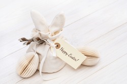 Easter bunny shaped Ivory gift bag with sweets and a gift tag with Happy Easter text and paper craft Easter egg on white wooden table . Willow branch