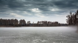 Overcast landscape on frozen misty lake  and moody sky in period between spring and winter
