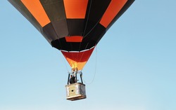 One flying striped black and orange big hot air balloon with people in a basket against a clear blue sky on background.