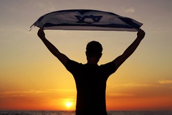 Israel, Israeli patriot, almost silhouette, with the flag of Israel over his head stands on the shores of the Mediterranean Sea at sunset. Independence day Israel, patriotic holiday concept