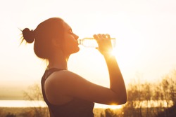 Beautiful girl in sportswear drinks water from a bottle at sunset
