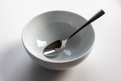 Empty white porcelain bowl with a polished silver spoon on a white background shows breakfast preparation without money for food and nutrition like cornflakes and others as clean setup on clean desk