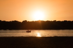 A yacht cruising along the river with a male sitting at the front of it, enjoying the picturesque sunset.