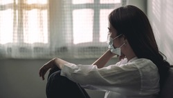 Close-up of an upset Asian patient woman wearing a face mask at home or hospital alone on a sofa waiting for a doctor. Hospital and Health care during Coronavirus or Covid-19 quarantine concept.
