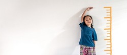 An adorable Asian little child girl measuring the height growth with the orange drawing on the white wall background. Girl power future and dream concept idea. Copy space.