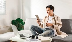 Happy smiling Asian student sits and relaxes on a couch using modern tablet browsing unlimited wireless internet, positive young man freelancer working uses wifi at a home apartment, technology idea.