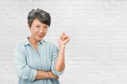 Beautiful old Asian woman with gray hair, wearing glasses and blue shirt, over white brick wall background. Copy space text for backdrop, artwork design. Elegant happy elderly retire concept.