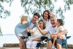Group of Multi generation Asian family using digital tablet video call with their family friend on the beach in summer day. Happy big family enjoy and having fun together in summer holiday vacation