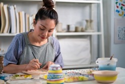 Asian woman learning color painting her self-made pottery at home. Confidence female enjoy hobbies and indoors leisure activity handicraft ceramic sculpture and painting workshop at pottery studio.