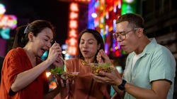 Group of Asian woman and LGBTQ people friends tourist enjoy eating traditional street food bbq seafood grilled squid with spicy sauce together at china town street night market in Bangkok, Thailand