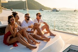 Group of man and woman friends enjoy party drinking champagne with talking together while catamaran boat sailing at summer sunset. Male and female relax outdoor lifestyle on tropical travel vacation