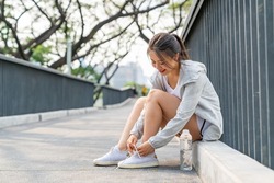 Asian woman in sportswear tying running shoelaces while jogging at public park in the city in summer morning. Healthy female athlete enjoy outdoor lifestyle sport training workout running exercise