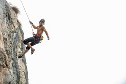 Confidence young Asian man climber with safety rope climbing on rocky mountain at tropical island in sunny day. Strong handsome male enjoy outdoor active lifestyle and extreme sport on summer vacation