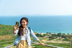 Young Asian woman traveler using mobile phone taking selfie while solo travel on tropical island mountain in summer sunny day. Cheerful female enjoy outdoor lifestyle in holiday beach vacation trip