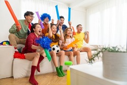 Group of Asian people friends sit on sofa watching and cheering soccer games competition on TV together at home. Happy man and woman sport fans shouting and celebrating sport team victory sports match