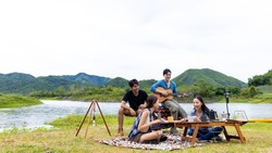 Group of Asian man and woman friends singing and playing guitar during having breakfast by the lake together. People enjoy and fun outdoor lifestyle travel nature camping on summer holiday vacation.