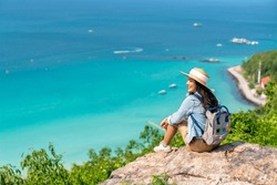Asian woman with backpack travel at tropical island and resting on mountain peak in summer sunny day. Attractive girl enjoy outdoor lifestyle looking beautiful ocean nature on beach holiday vacation.