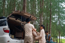 Group of Asian people friends enjoy outdoor lifestyle road trip and camping together on summer holiday travel vacation. Man and woman taking off camping supplies from car trunk at natural park.