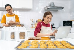 Asian man and woman bakery shop owner working on laptop computer and listing customer order in kitchen. Bakery chef making cookies for selling online delivery. Small business food and drink concept.