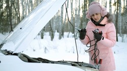 Young Woman in Warm Clothes Trying to Fix Discharged Battery of her Car with Booster Jumper Cables in Cold Winter Day