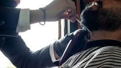 Hands of a barber attending to a customer cutting his beard using a clipper and comb, both are unrecognizable. Lifestyle.