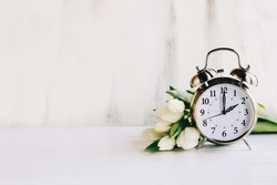 Daylight savings time concept. Set your clocks and to 2 am and spring ahead with this image of an alarm clock with white tulip flowers. Selective focus with blurred foreground and background.
