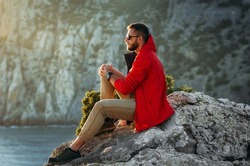 Portrait of a brutal bearded man in a red jacket by the sea. Male portrait on the background of a seascape. A traveler on the background of the sea coast. A man in a jacket at sunset