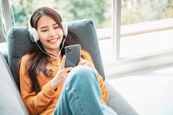 Happy asian woman using mobile smartphone and headphones while listening online music media entertainment relaxation in a sofa at home. copy space.