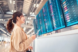 Happy asian woman traveller checking flight schedule departures board in airport terminal hall in front of check in counters. Tourist journey trip concept