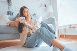 Happy young asian woman drinking coffee relaxing on the rug beside to the sofa at home. Smiling female enjoying resting sitting on couch in modern living room.
