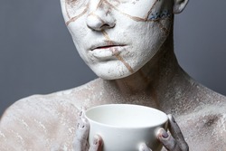 Art portrait of woman covered in clay isolated over grey background. Woman face like cracked earth holding  a cup