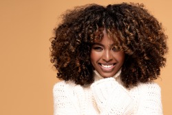 Happy beautiful african girl with afro hairstyle posing in cozy sweater on beige studio background.
