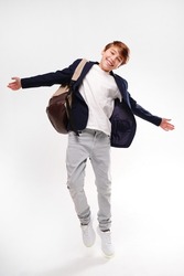 Young energetic handsome happy school boy in fashionable clothes jumping with backpack over studio background, smiling to the camera. Real people emotions. Full photo.