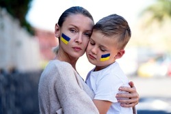 Families against war. Upset Ukrainian boy with his mother, with bue yellow flag on his face,  protesting war conflict. Evacuation of civilians. Freedom to Ukraine. Stop Russian aggression. Stop war 