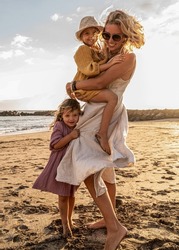 Happy beautiful family relaxing and walking at the sunset beach in summer. Smiling mother with her two little daughters having fun on vacations. Real people lifestyle. 
