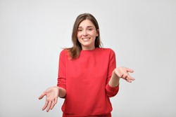 Studio portrait of attractive caucasian teenager in red pulover shrugging her shoulders having some doubts. Emotional woman confused while making some decisions on gray background