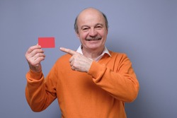 Happy senior hispanic bald man advertising bonus card pointing on it with finger smiling at camera. Guy going to pay by credit card, doing shopping