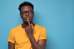 Young african american man, student with finger in mouth in deep thought, isolated on blue background. Negative emotion, facial expression, feelings