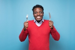 Hungry african young man holding fork and knife on hand ready to eat being excited.Diet and healthy food concept.