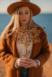 A woman walking along the coast near the sea. An elegant lady in a brown coat and a hat with fashionable makeup walks on the seashore